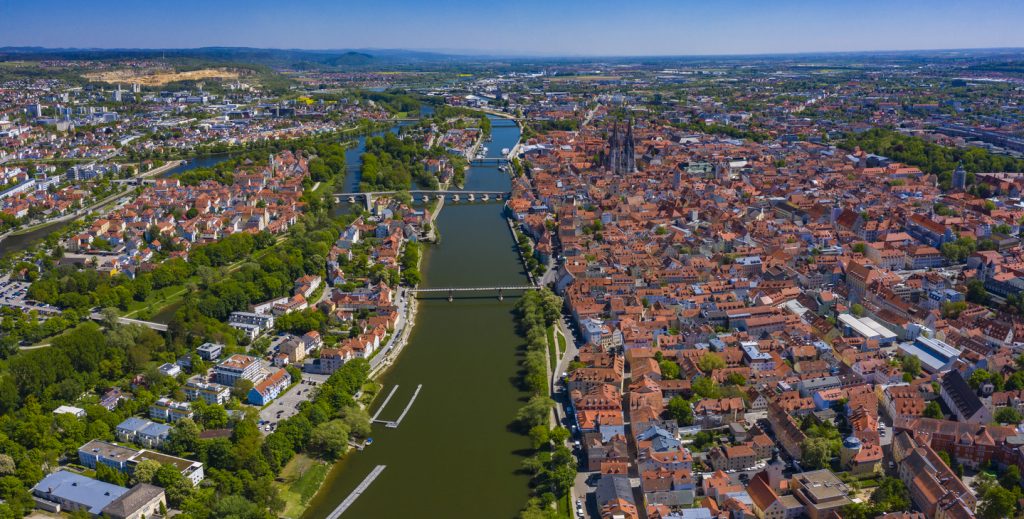 Panorama von Regensburg mit Domtürmen, roten Dächern und der Donau im Vordergrund.