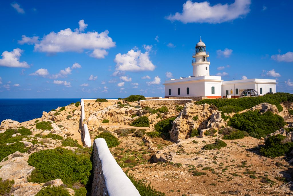 Der weiße Leuchtturm von Cavalleria steht auf einer felsigen Küste mit Blick auf das blaue Meer.