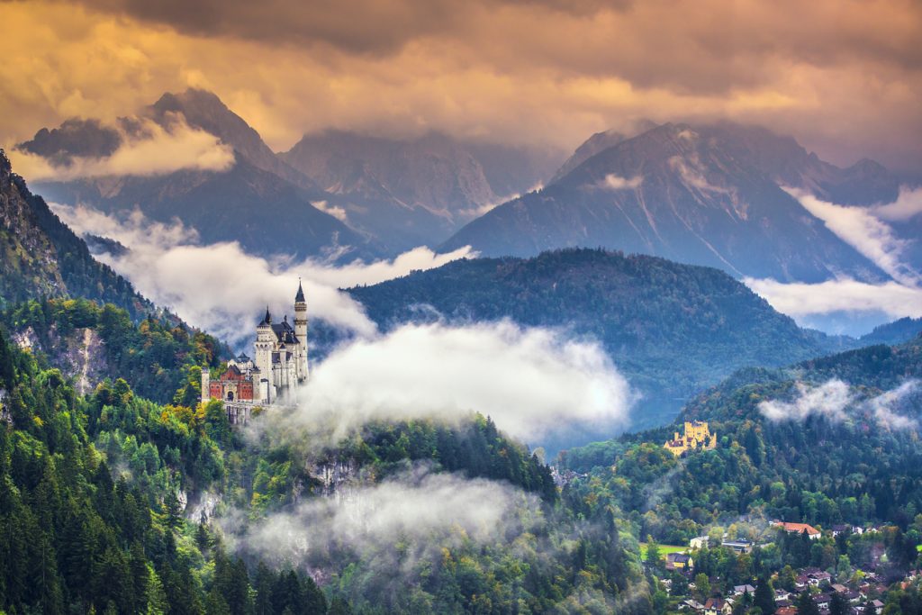 Schloss Neuschwanstein ragt aus Wolken und Wald vor dramatischer Bergkulisse hervor.