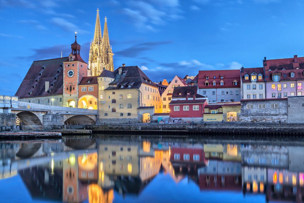 Beleuchtete Häuserfronten in Regensburg spiegeln sich im ruhigen Wasser vor dem Dom.