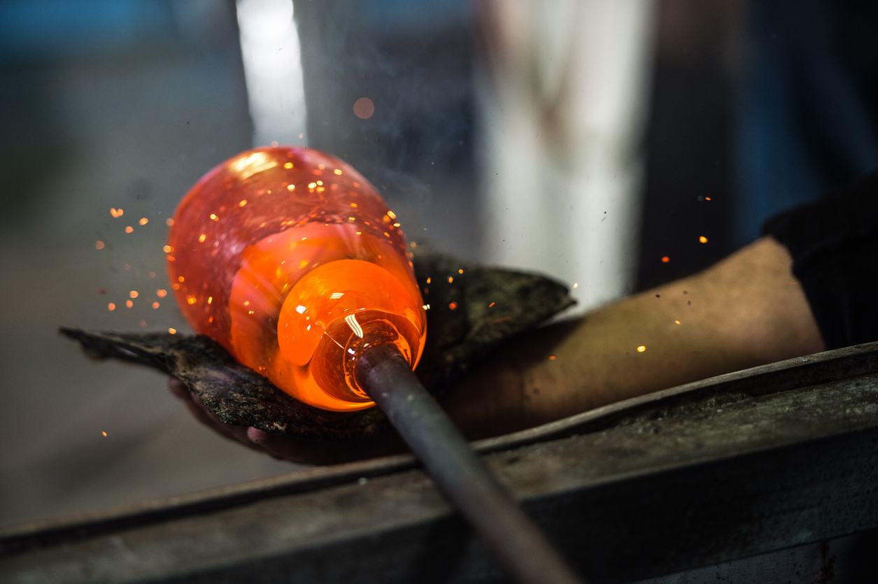 Nahaufnahme aus einer Glasbläserei, in der eine orange glühende Glasblase mit Werkzeugen auf der Werkbank geformt wird.