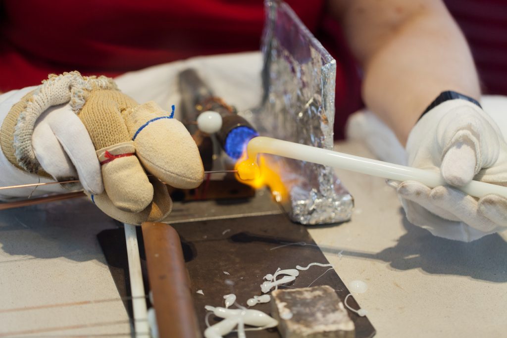 Mit Handschuhen wird ein Stab am Brenner erhitzt und geformt, während das Material am Ende glüht.