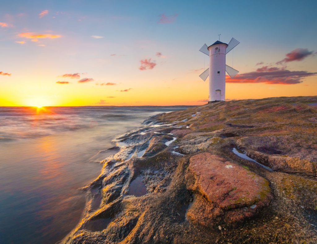 Eine weiße Windmühle steht auf Felsen am Wasser, während die Sonne am Horizont untergeht.