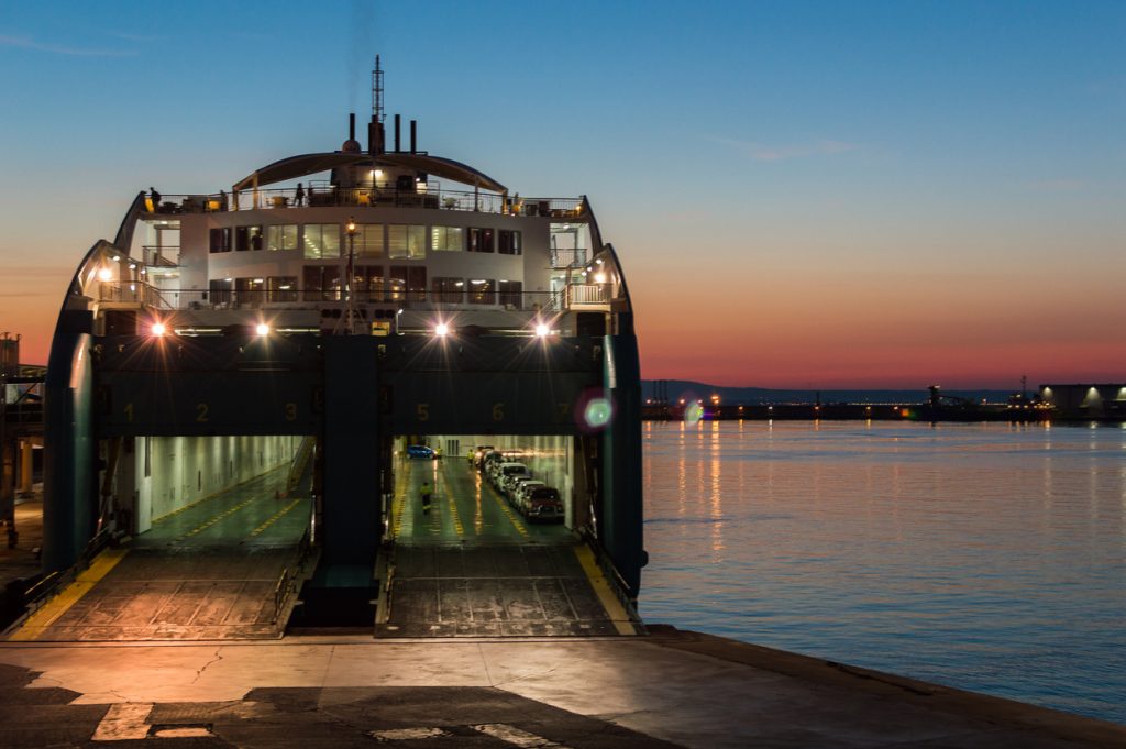 Eine Fähre liegt bei Sonnenuntergang im Hafen, die Heckklappe ist geöffnet und Autos stehen an Bord.