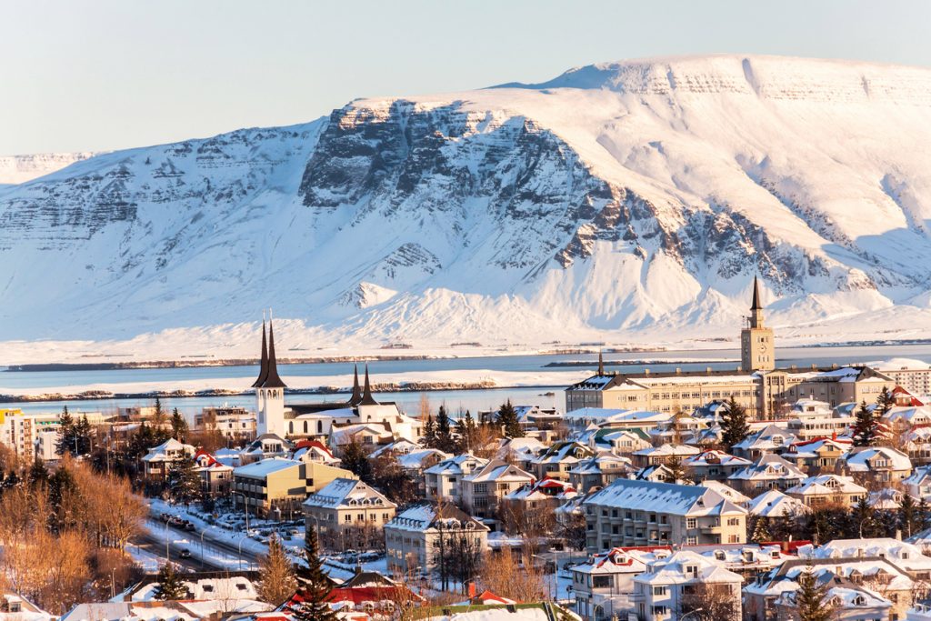 Verschneite Häuser in Reykjavik mit Kirchturmspitzen, Hafen und schneebedecktem Bergmassiv dahinter.
