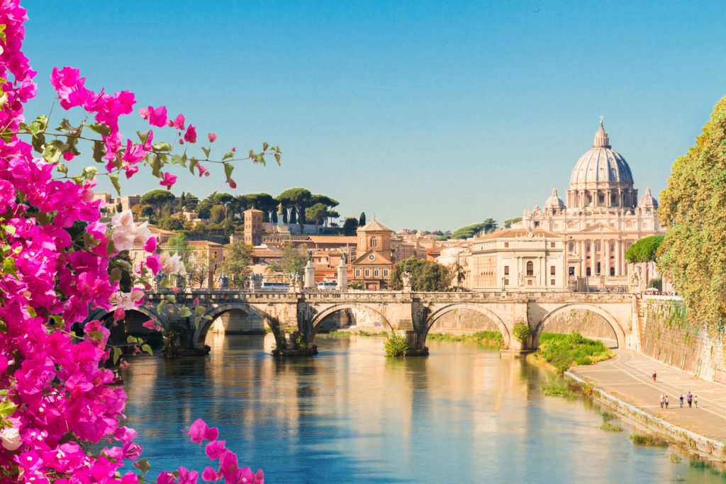 Blick über den Tiber mit Steinbrücke und Petersdom im Hintergrund, eingerahmt von pinken Bougainvillea-Blüten.