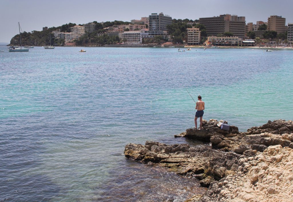 Ein Mann angelt von Felsen aus in ruhigem Meer, im Hintergrund liegen Hotels an der Küste.