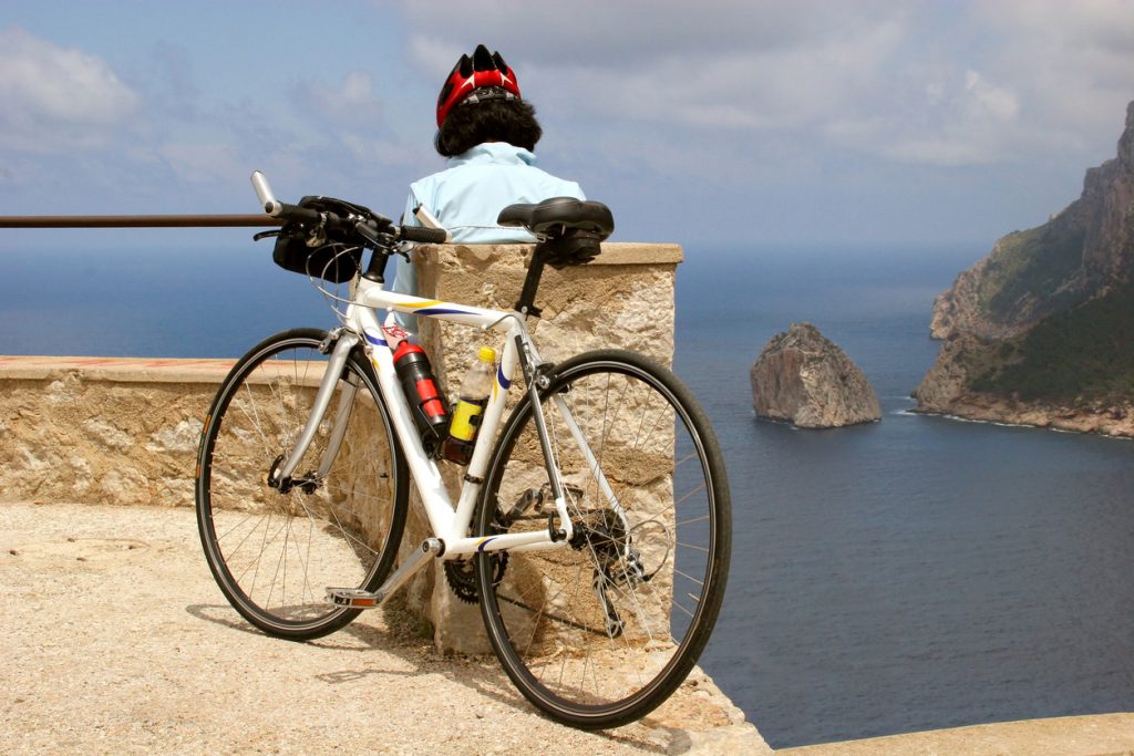 Ein Rennrad lehnt an einer Steinmauer, während eine Person mit Helm auf das Meer und die Felsen blickt.