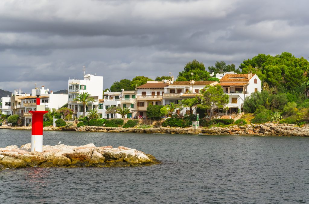 Ein rot weißes Leuchtfeuer steht auf Felsen vor einer Uferpromenade mit Häusern am Wasser.
