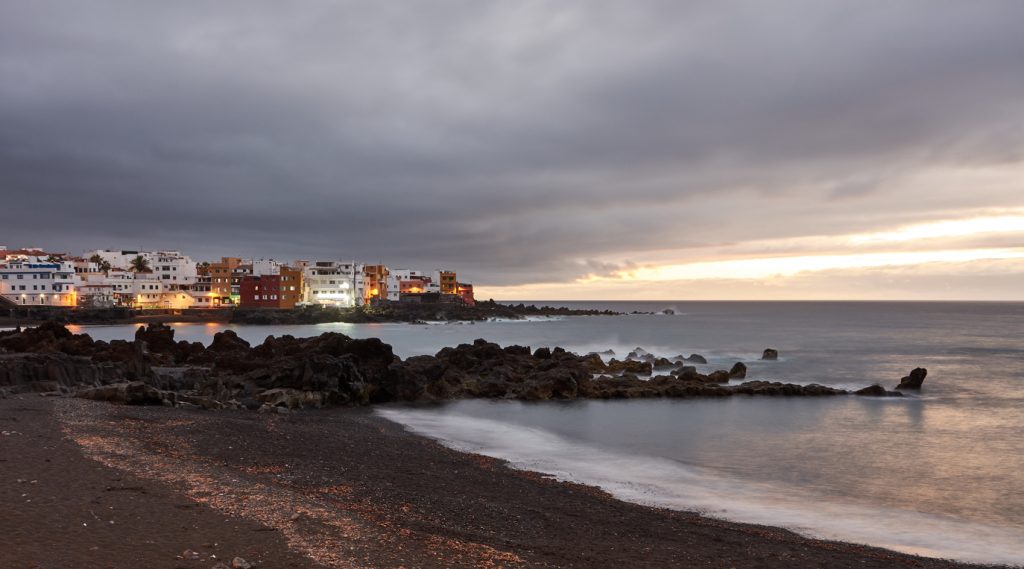 Dunkler Lavastrand der Playa Jardín mit Brandung, Felsen und beleuchteten Häusern am Horizont.