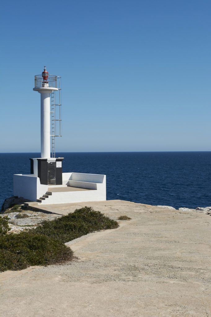 Ein schlanker weißer Leuchtturm steht auf einer Plattform am Meer vor tiefblauem Wasser und klarem Himmel.