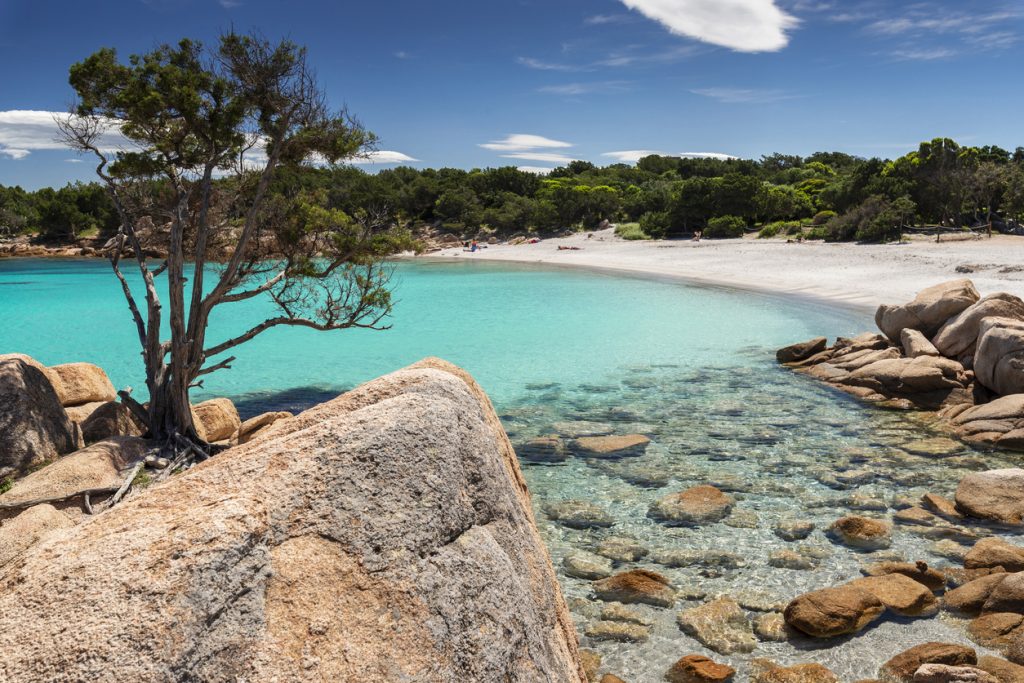 In einer geschützten Bucht liegt helles Wasser zwischen großen Granitfelsen und einem kleinen Strand.