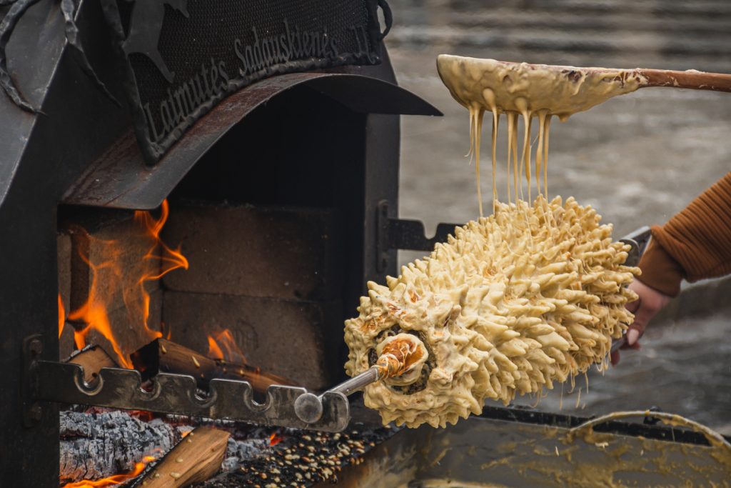 Ein Baumkuchen wird an einem Spieß über offenem Feuer gebacken, während Teig in Fäden herunterläuft.