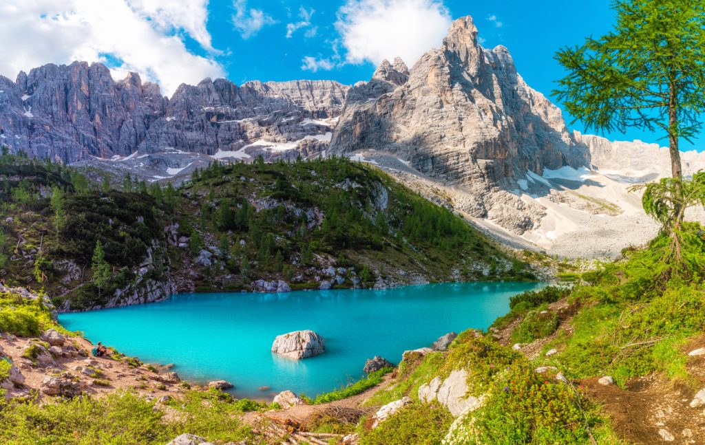 Ein leuchtend türkiser Bergsee liegt in einem Felskessel, umgeben von steilen grauen Dolomiten-Wänden und grünen Hängen.