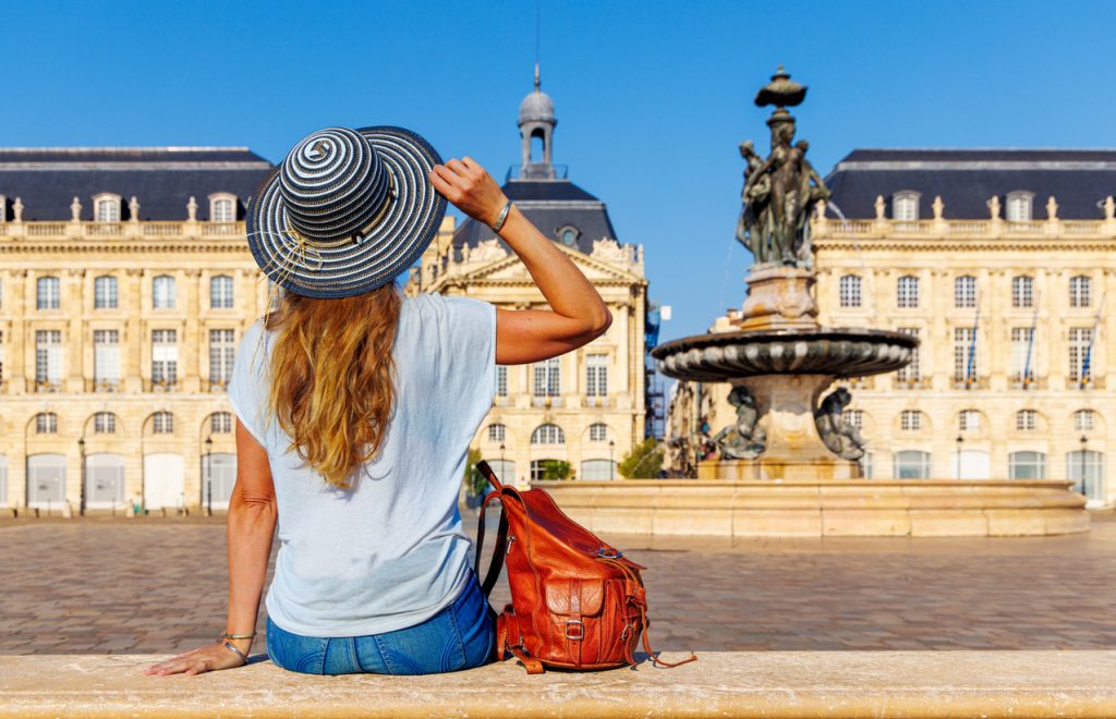 Frau mit Hut sitzt vor einem Brunnen auf einem großen Platz mit historischen Gebäuden