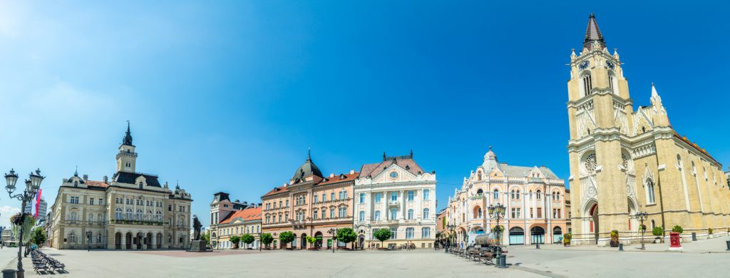 Panorama des Freiheitsplatzes in Novi Sad mit Rathaus und hoher Kirche unter blauem Himmel.