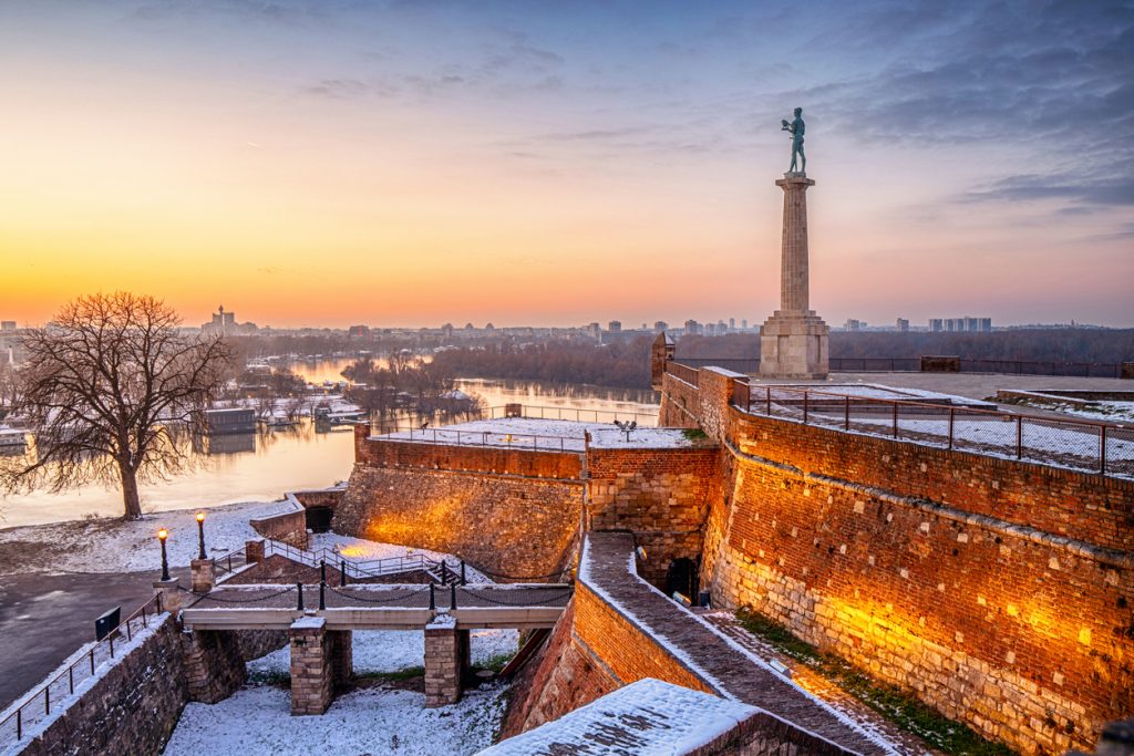 Festungsmauer mit Statue auf hoher Säule bei winterlichem Sonnenuntergang und Flussblick.