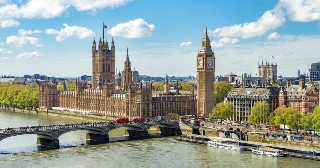 Die Houses of Parliament mit dem Elizabeth Tower am Flussufer bei blauem Himmel.