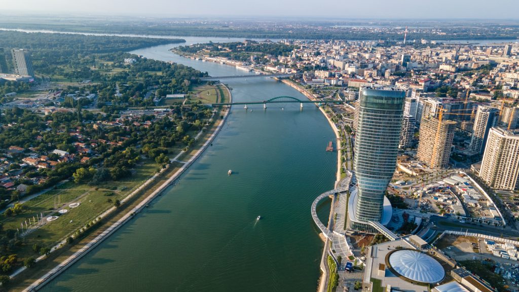 Luftaufnahme eines Flusses mit Hochhaus und Brücke, dahinter dichte Stadtbebauung.