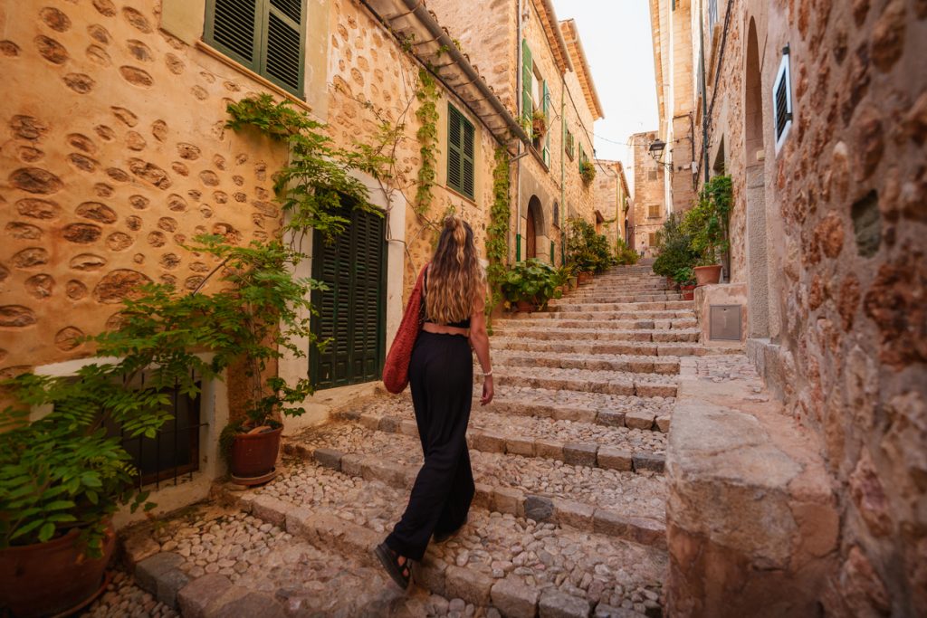 Frau läuft eine steinerne Treppe durch eine schmale Gasse mit grünen Fensterläden in Fornalutx hinauf.