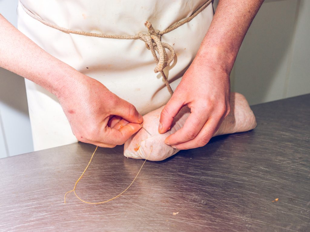 Hände binden eine gefüllte Wursthülle auf einer Edelstahlfläche fest.