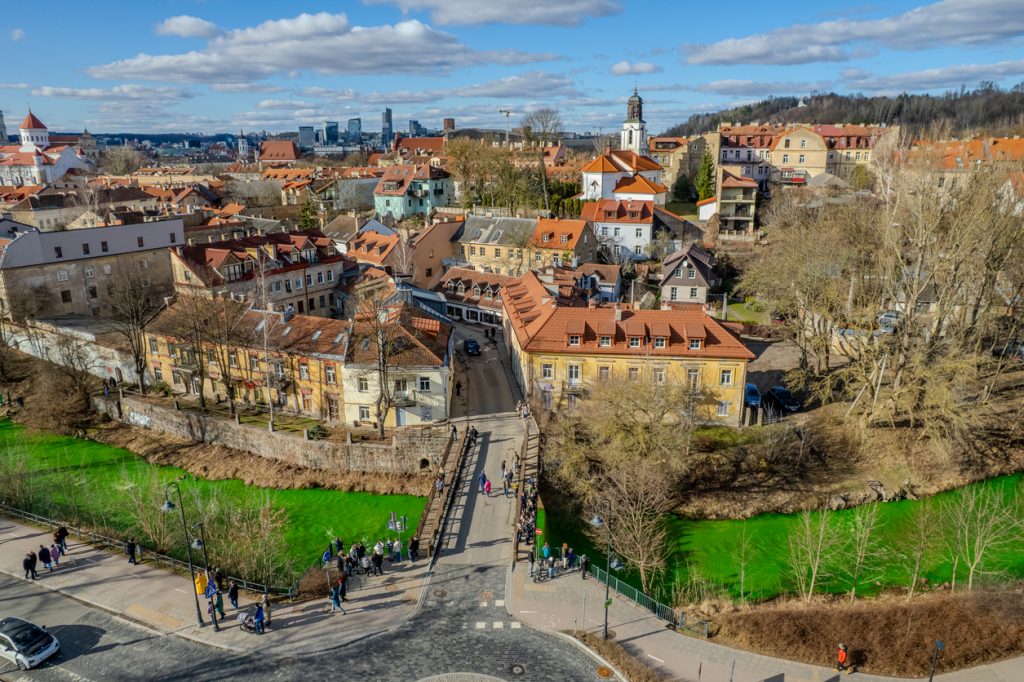 Eine Brücke führt in ein Viertel mit roten Dächern und Kirchen, daneben fließt ein auffällig grüner Fluss.
