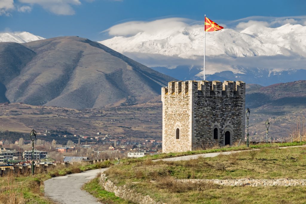 Ein steinerner Turm mit Flagge steht in einer weiten Landschaft vor schneebedeckten Bergen nahe Skopje.