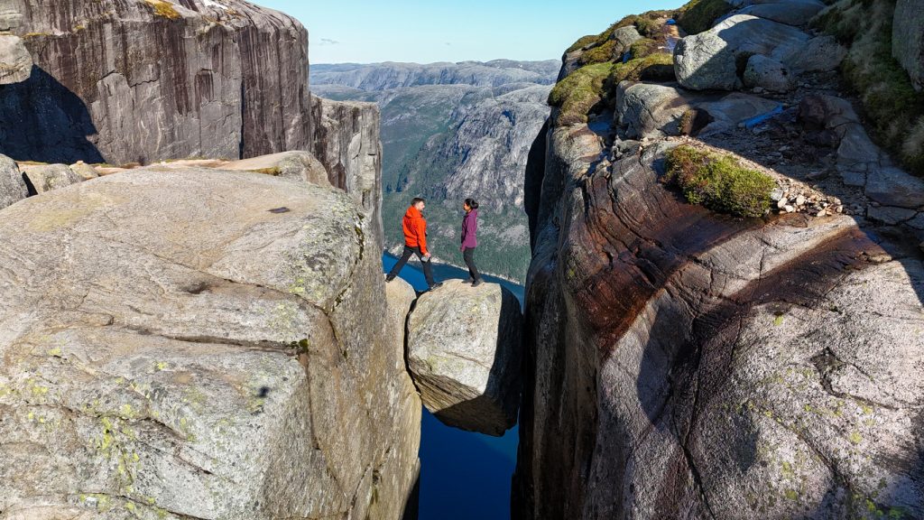 Zwei Personen stehen auf einem eingeklemmten Felsblock hoch über einem Fjord.