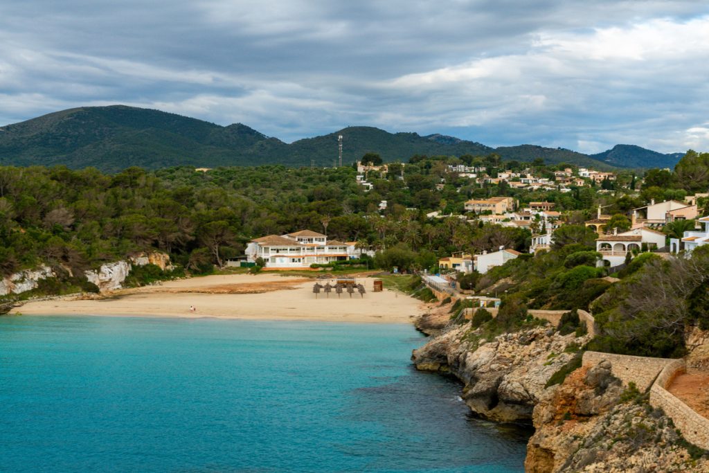 Eine kleine Sandbucht mit türkisblauem Wasser, Häusern am Hang und Bergen im Hintergrund in Cala Romàntica.