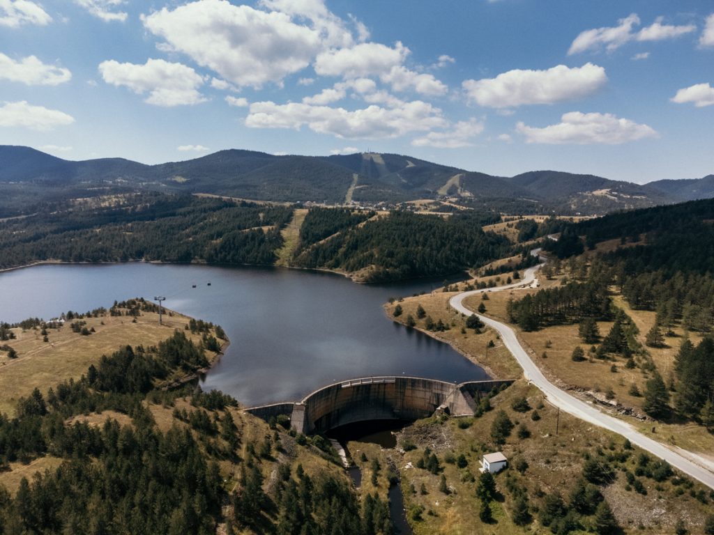 Ein See mit einer Staumauer liegt zwischen bewaldeten Hügeln, daneben führt eine Straße durch die Landschaft.