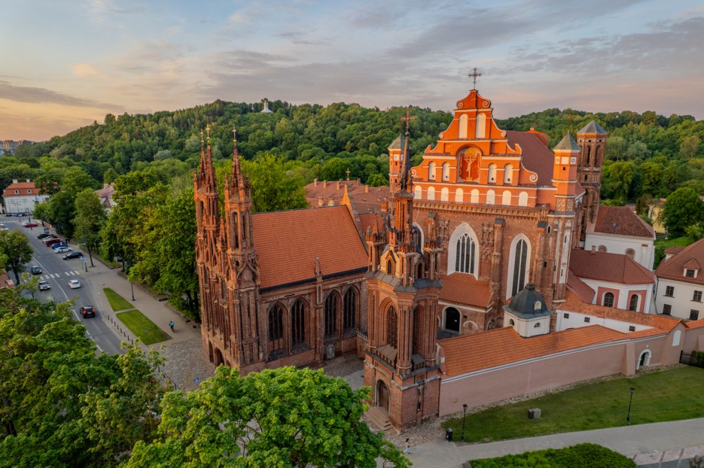 Eine rote Backsteinkirche und historische Gebäude leuchten vor einem grünen Hügel und ruhigem Himmel.
