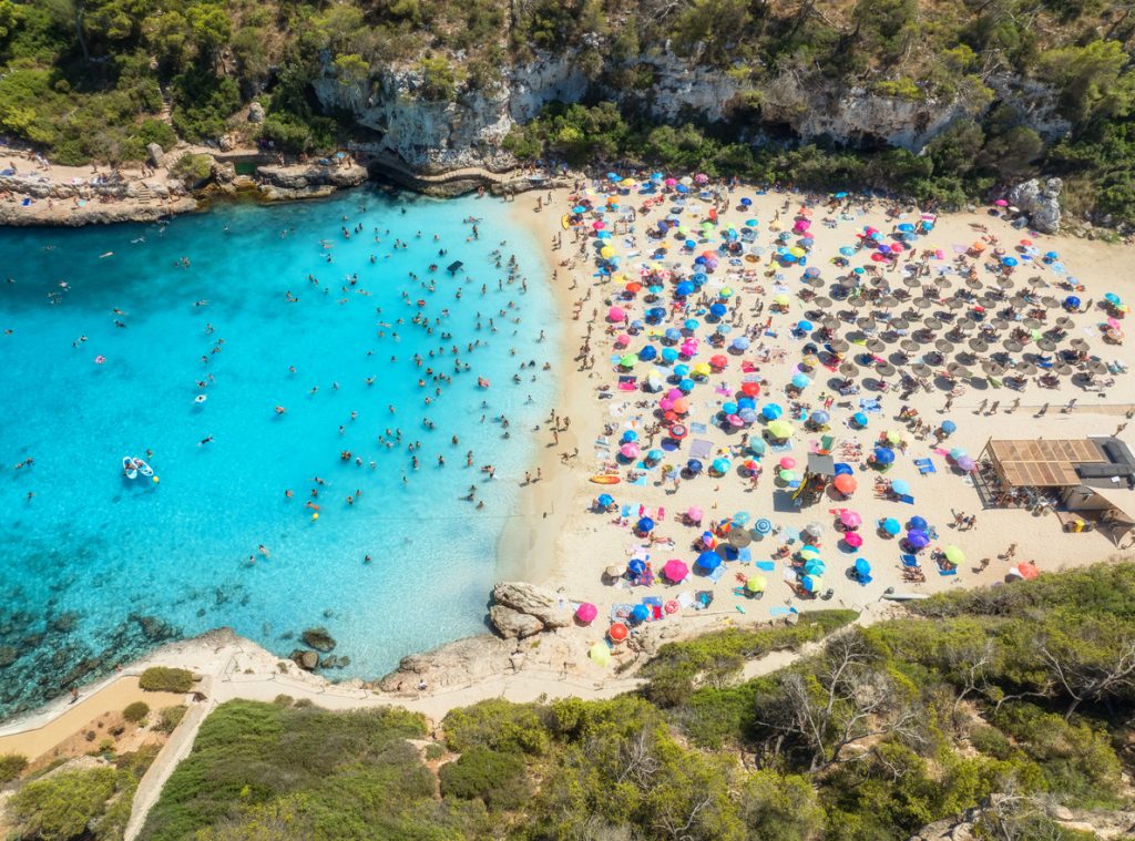 Luftaufnahme eines vollen Strandes mit vielen bunten Sonnenschirmen und leuchtend blauem Wasser in Cala Romàntica.