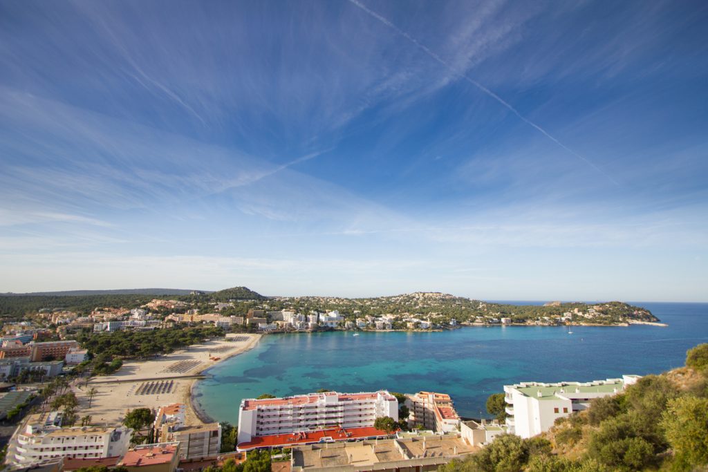 Eine breite Bucht mit Strand und Gebäuden liegt unter einem hohen Himmel mit feinen Wolkenspuren.