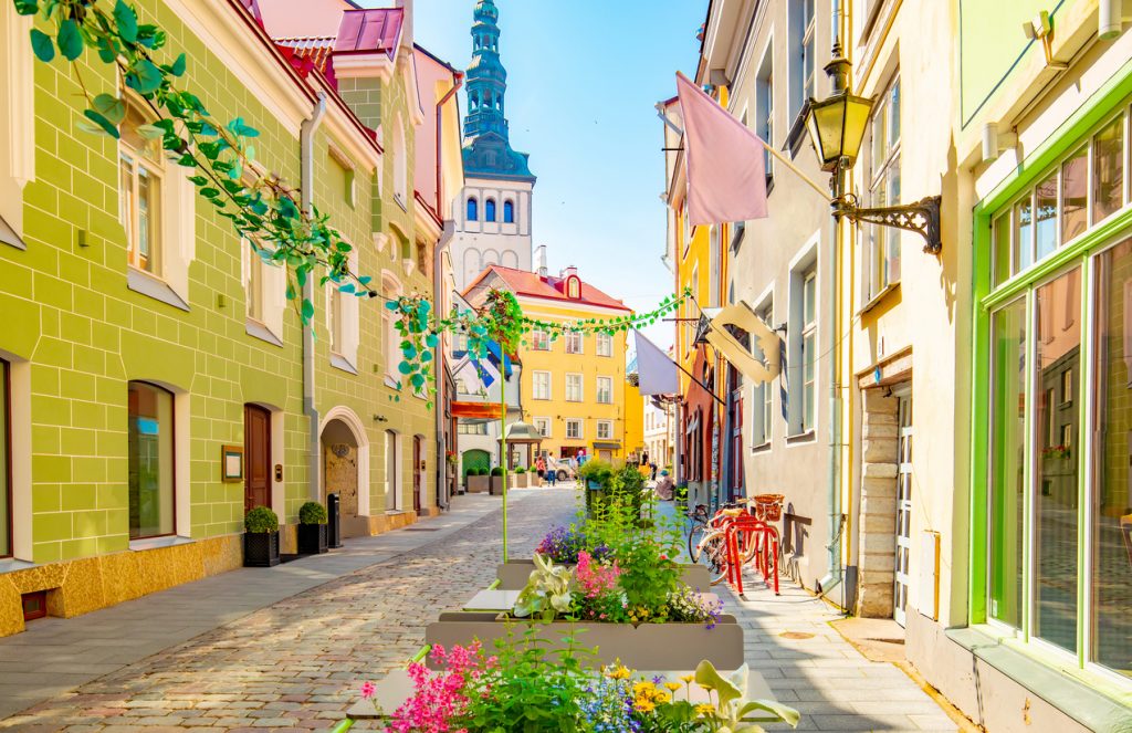 Schmale Straße mit pastellfarbenen Häusern, Blumenkästen und einem Turm in der Ferne.