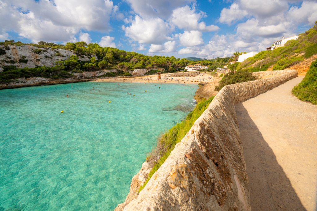 Ein heller Küstenweg läuft an einer türkisfarbenen Bucht entlang, im Hintergrund Strand und Häuser in Cala Romàntica.