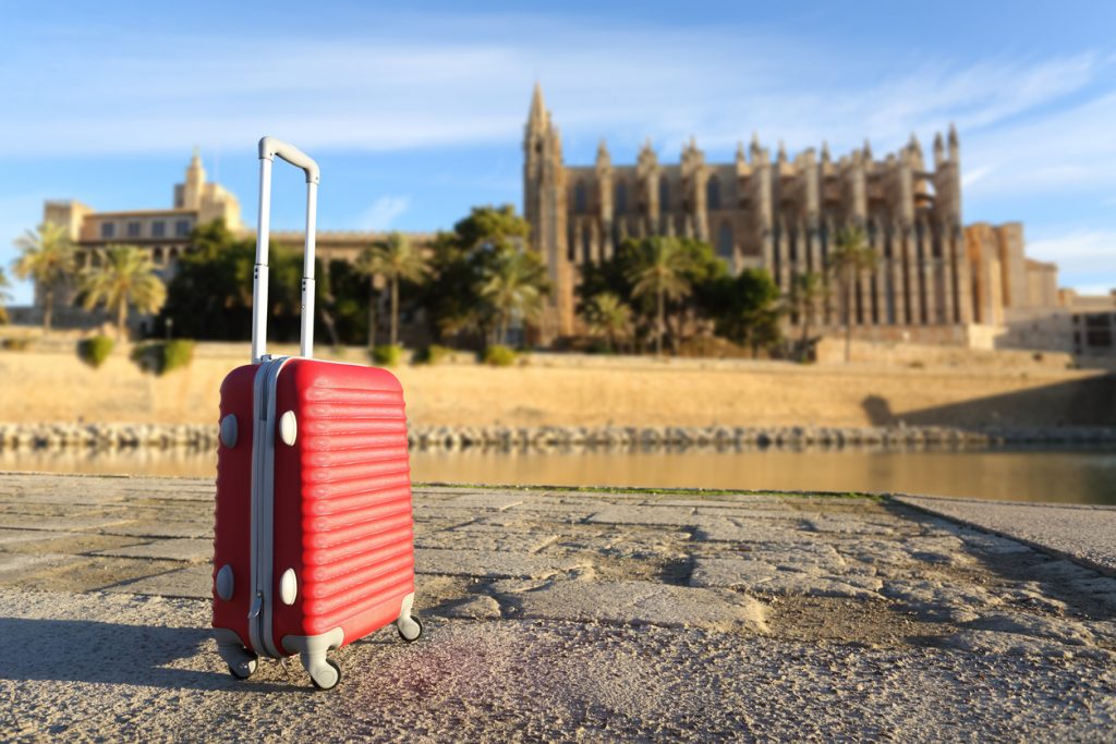 Ein roter Koffer steht in Mallorca am Wasser, im Hintergrund ist die Kathedrale von Palma unscharf zu sehen.