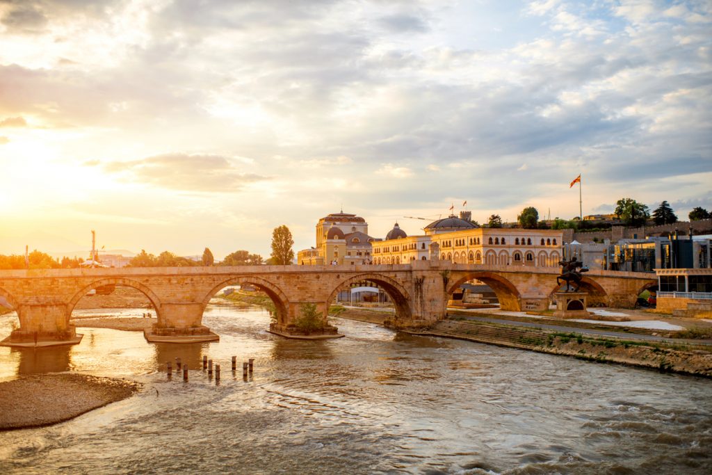 Eine alte Steinbrücke führt über den Fluss, während die Sonne warmes Licht auf Skopje wirft.