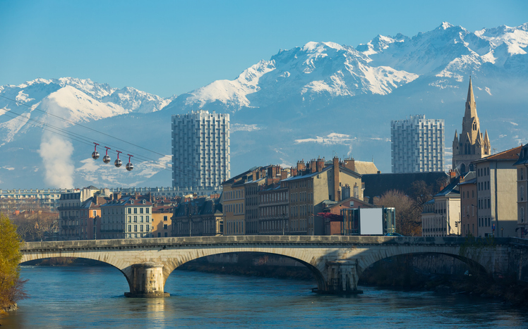 Blick über eine steinerne Bogenbrücke und historische Stadthäuser auf die gewaltigen, verschneiten Gipfel der Belledonne-Kette unter strahlend blauem Himmel.