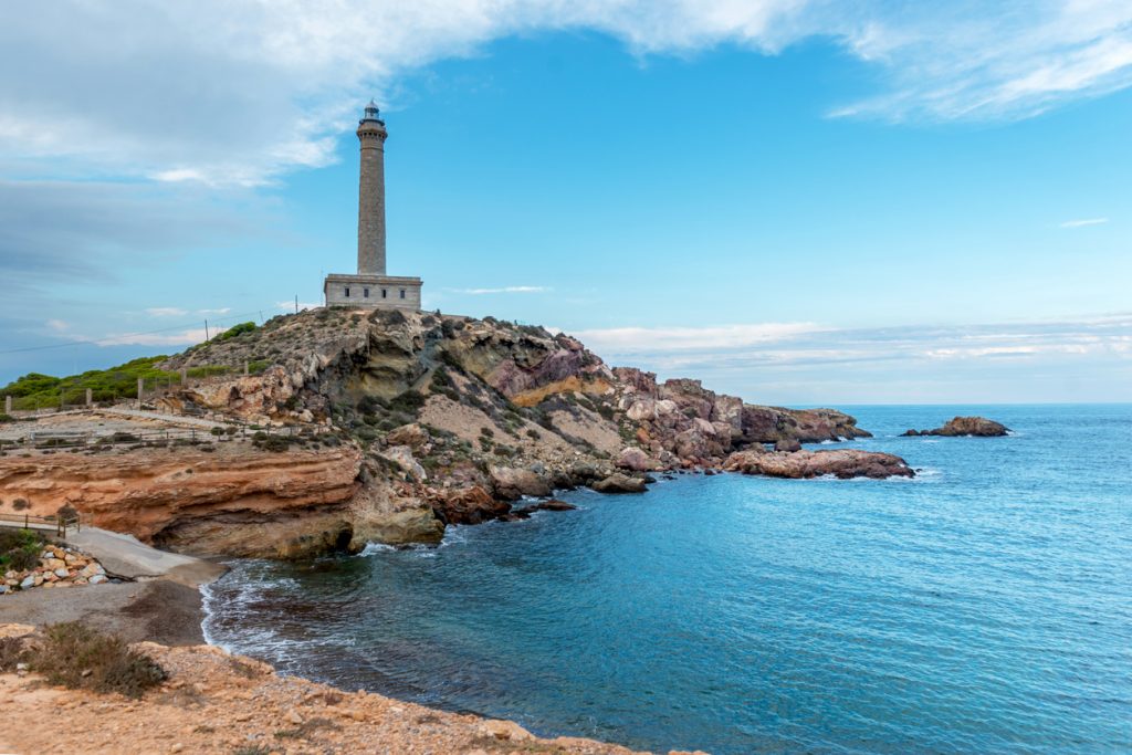 Ein hoher, steinerner Leuchtturm auf einer felsigen Landzunge unter blauem Himmel am Mittelmeer