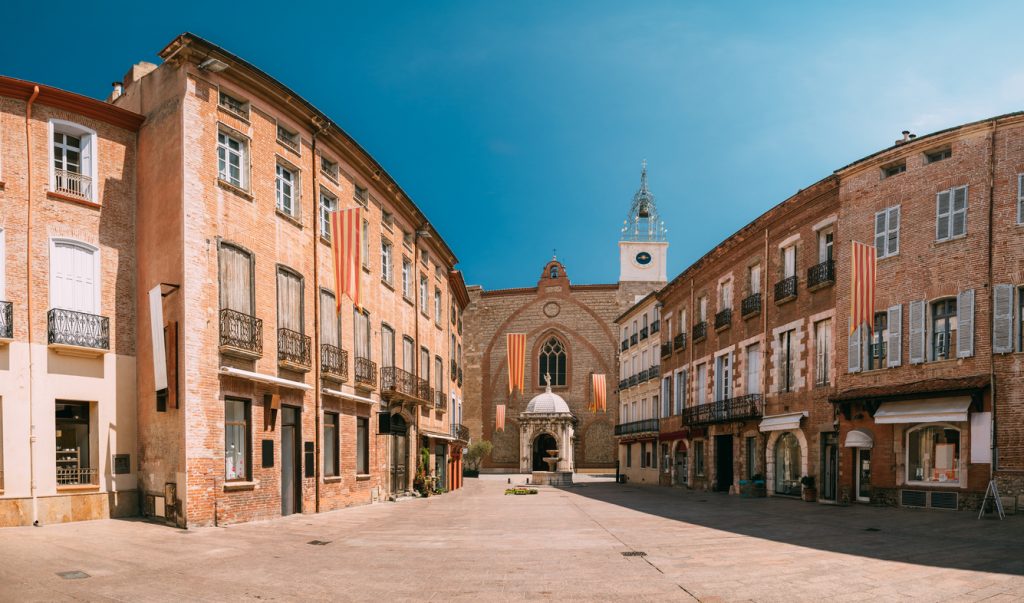 Weite Altstadtstraße mit Backsteinfassaden und Kirche im Hintergrund in Perpignan.