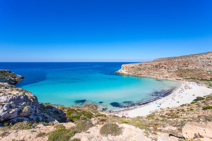 Ein sichelförmiger, weißer Sandstrand schmiegt sich an eine Bucht mit leuchtend türkisem Wasser unter blauem Himmel.