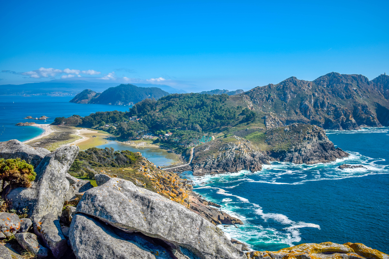 Ein spektakulärer Ausblick von einem felsigen Gipfel auf die bewaldeten Inseln, eine schmale Landzunge mit Sandstrand und das tiefblaue, schäumende Meer.