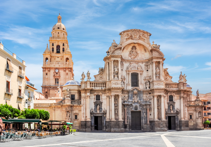 Die monumentale barocke Fassade der Kathedrale von Murcia mit ihren zahlreichen Statuen und Säulen steht an einem weiten Platz mit Außengastronomie.