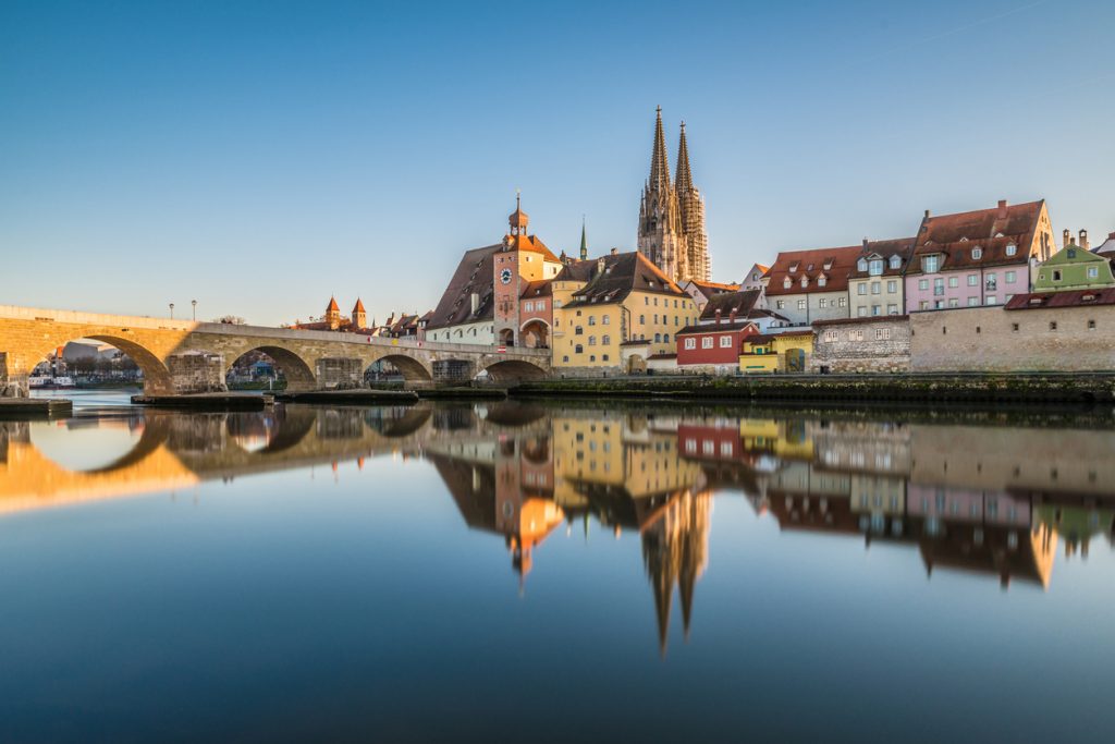 Die Steinerne Brücke und der Dom von Regensburg spiegeln sich bei Sonnenaufgang perfekt in der glatten Oberfläche der Donau.