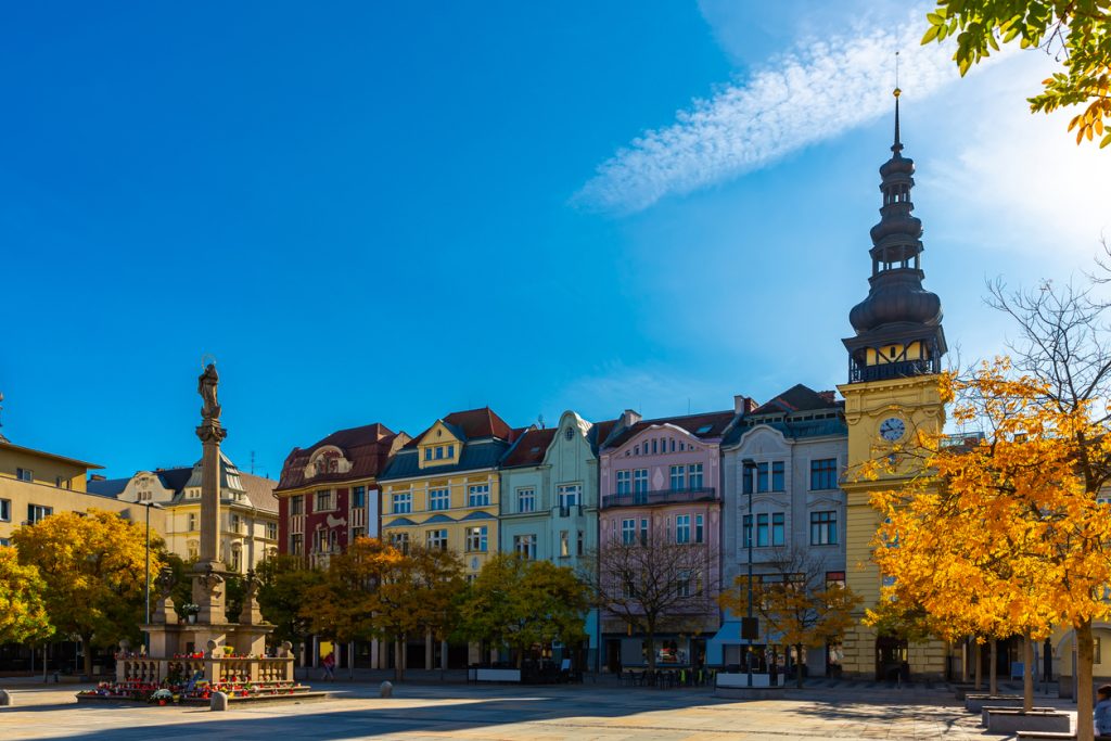 Weitläufiger Stadtplatz mit Denkmal, bunten Fassaden und Turm im Hintergrund