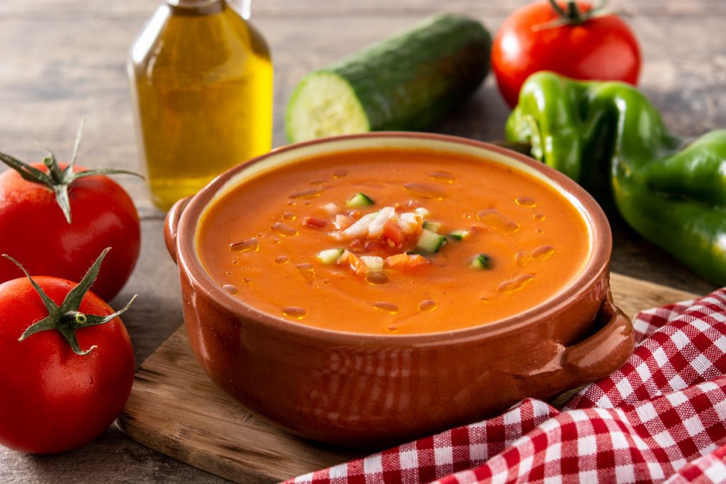 Eine rustikale Tonschale mit orangeroter Gazpacho steht neben frischen Tomaten, Gurken und Paprika.