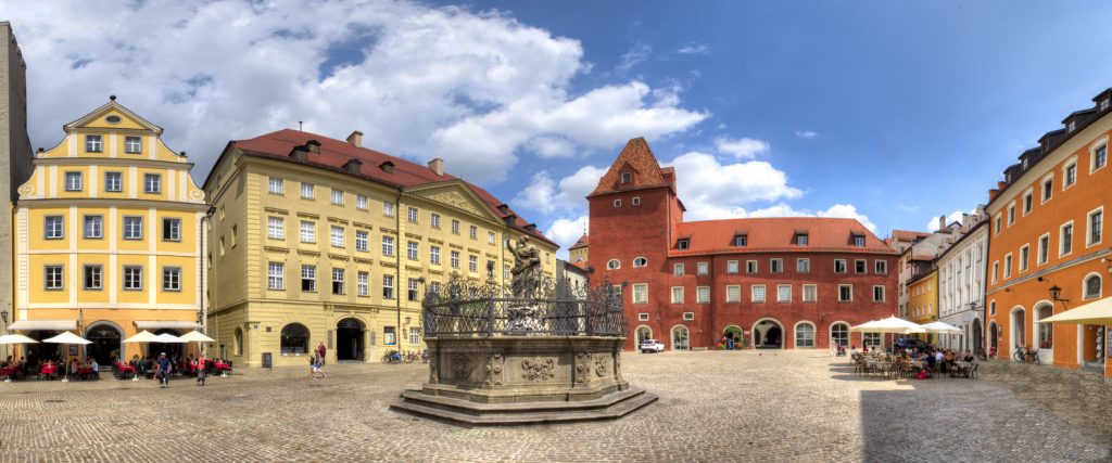 Ein weites Panorama des Haidplatzes in Regensburg mit farbenfrohen Patrizierhäusern, Straßencafés und dem zentralen Justitia-Brunnen.