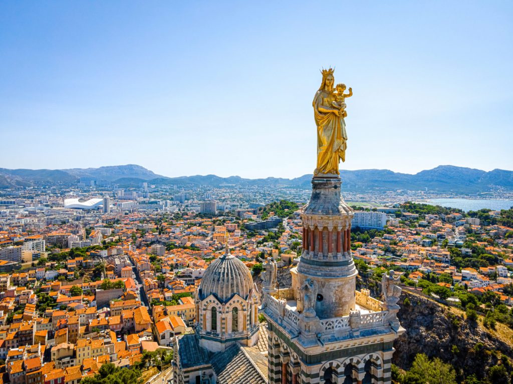 Eine goldene Marienstatue mit Kind krönt die Spitze eines Glockenturms, im Hintergrund erstreckt sich das weite Stadtpanorama bis zum Meer.