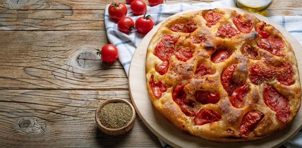 Eine runde, saftige Focaccia belegt mit vielen Kirschtomaten liegt auf einem Holzbrett; daneben stehen frische Tomaten und eine Schale mit getrockneten Kräutern.