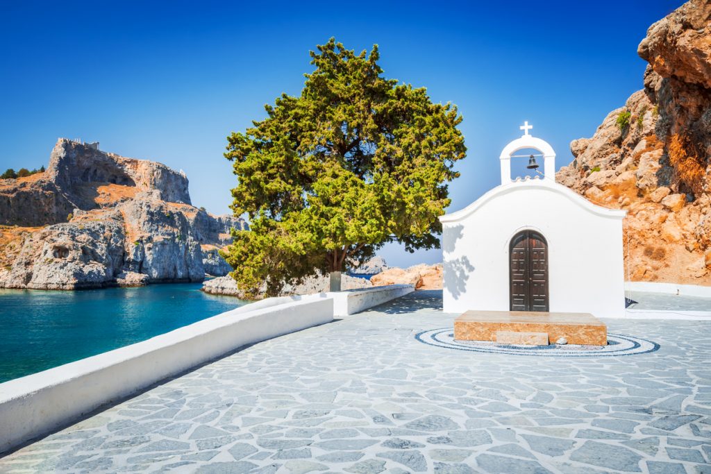 Eine strahlend weiße Kapelle mit kleiner Glocke und dunkler Holztür steht auf einem gepflasterten Vorplatz neben einem großen Baum; im Hintergrund ist eine schroffe Felsküste und das azurblaue Meer zu sehen.